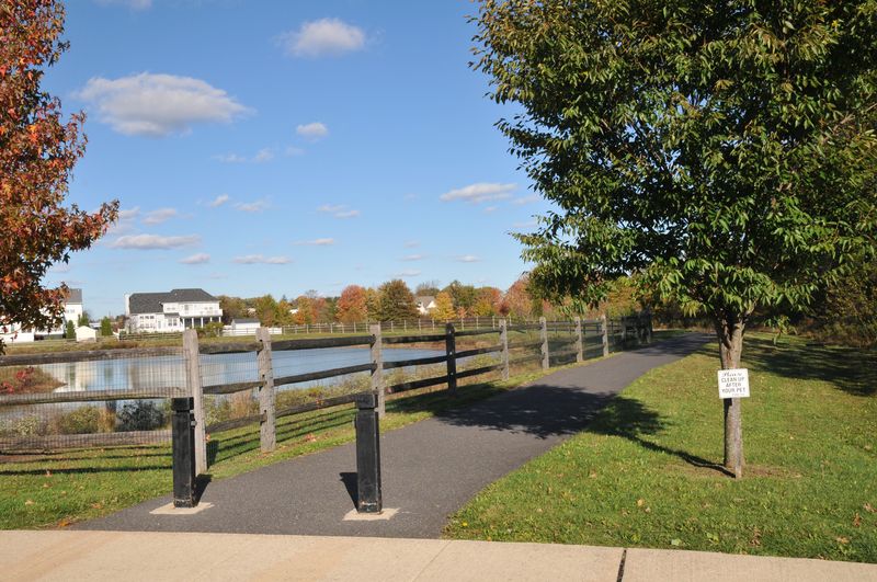 Paved walking path beside a pond with a split-rail fence