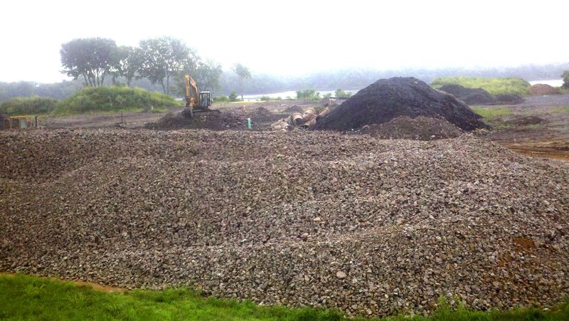 Site covered in rock with an excavator and soil piles behind